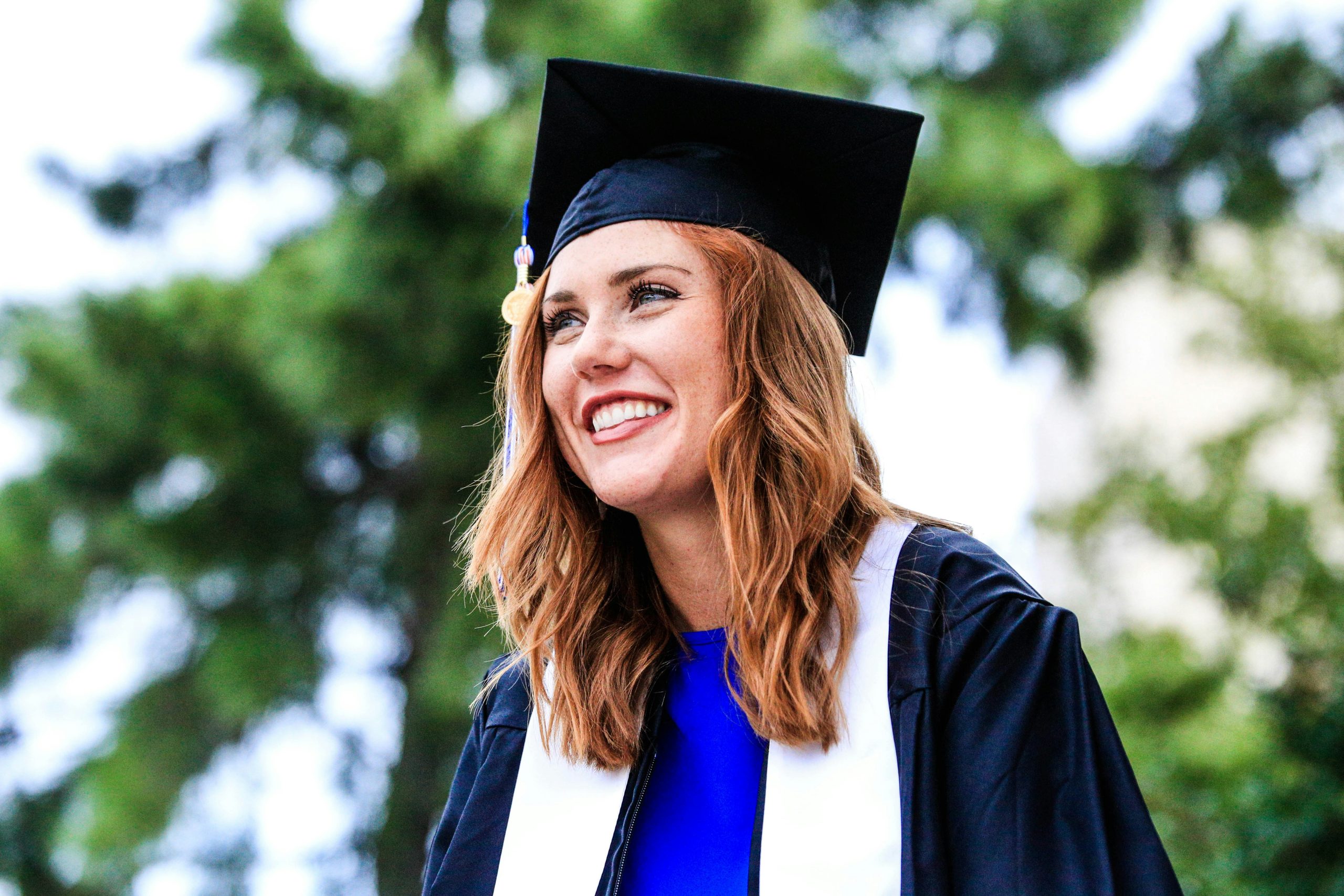 Vibrant outdoor portrait of a new graduate. She has a joyous smile and is wearing a graduation cap and gown.